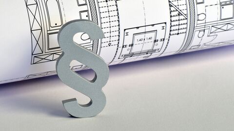 Bauplan mit Paragraph Ein zusammengerollter schwarz-weiß gezeichneter Bauplan liegt auf einem hellgrauen Tisch. Ein graues Paragraphenzeichen steht angelehnt an dem gerollten Plan im Vordergrund.