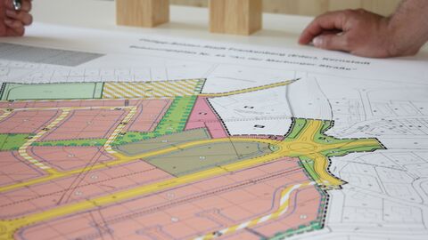 Bebauungsplan auf Tisch Ein Bebauungsplan mit verschiedenen eingezeichneten rosa, orange und grünen Flächen liegt auf einem Tisch und eine Person stützt ihre Hand neben dem Plan auf den Tisch.